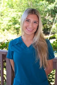 Young woman smiling at camera wearing blue polo shirt outdoors with green foliage background.